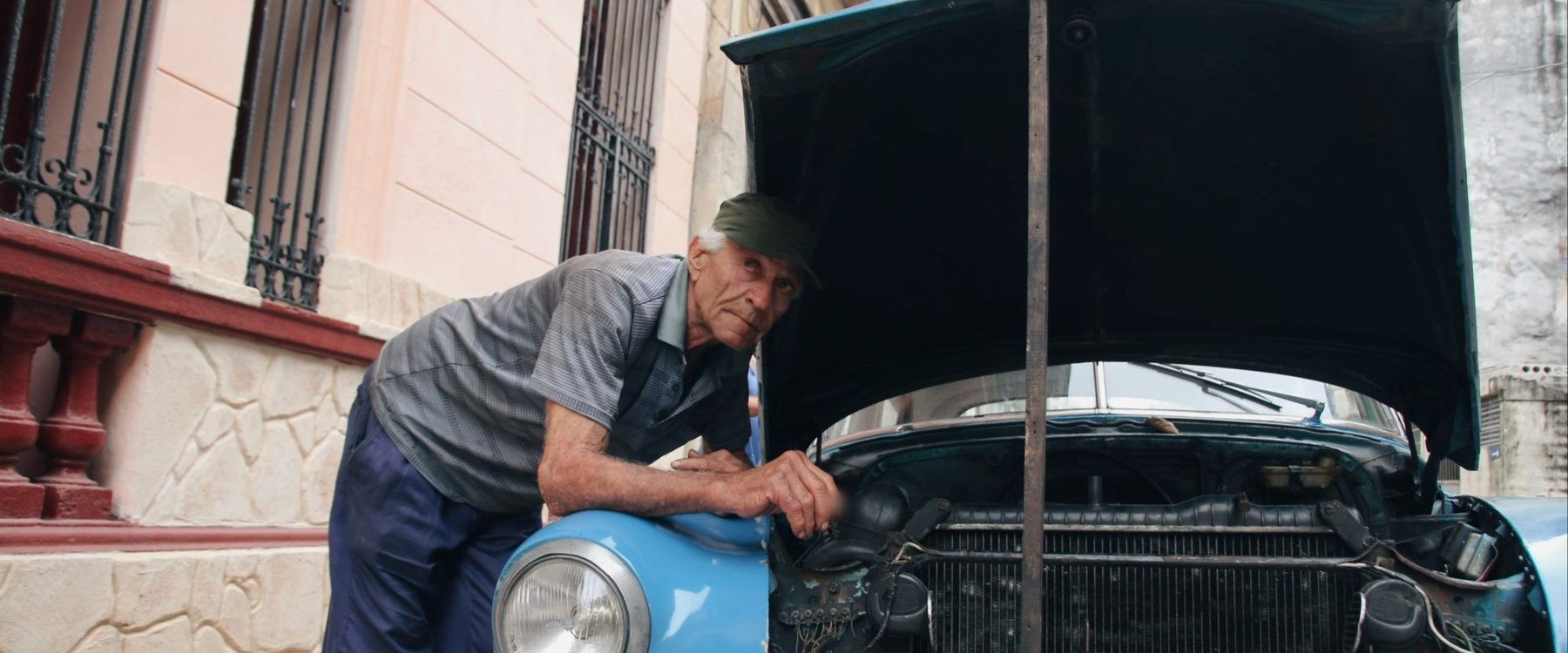 Mecânico de boné debruçado sobre o motor de um carro antigo azul, com o capô aberto, numa rua de calçada estreita
