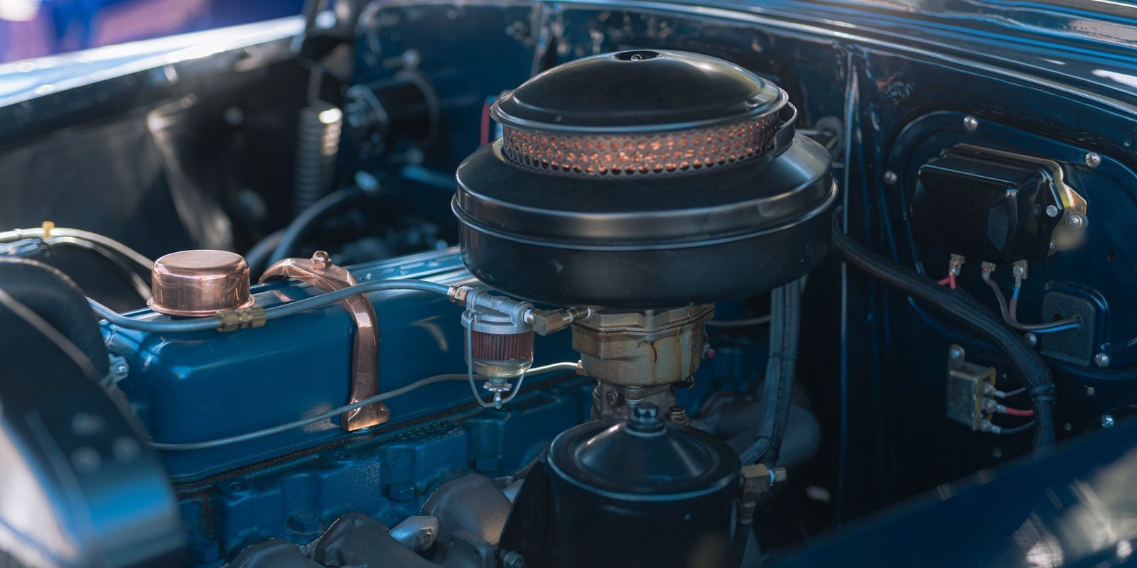 Compartimento do motor de um carro antigo pintado de azul, com tampa de óleo de cobre e filtro de ar em primeiro plano