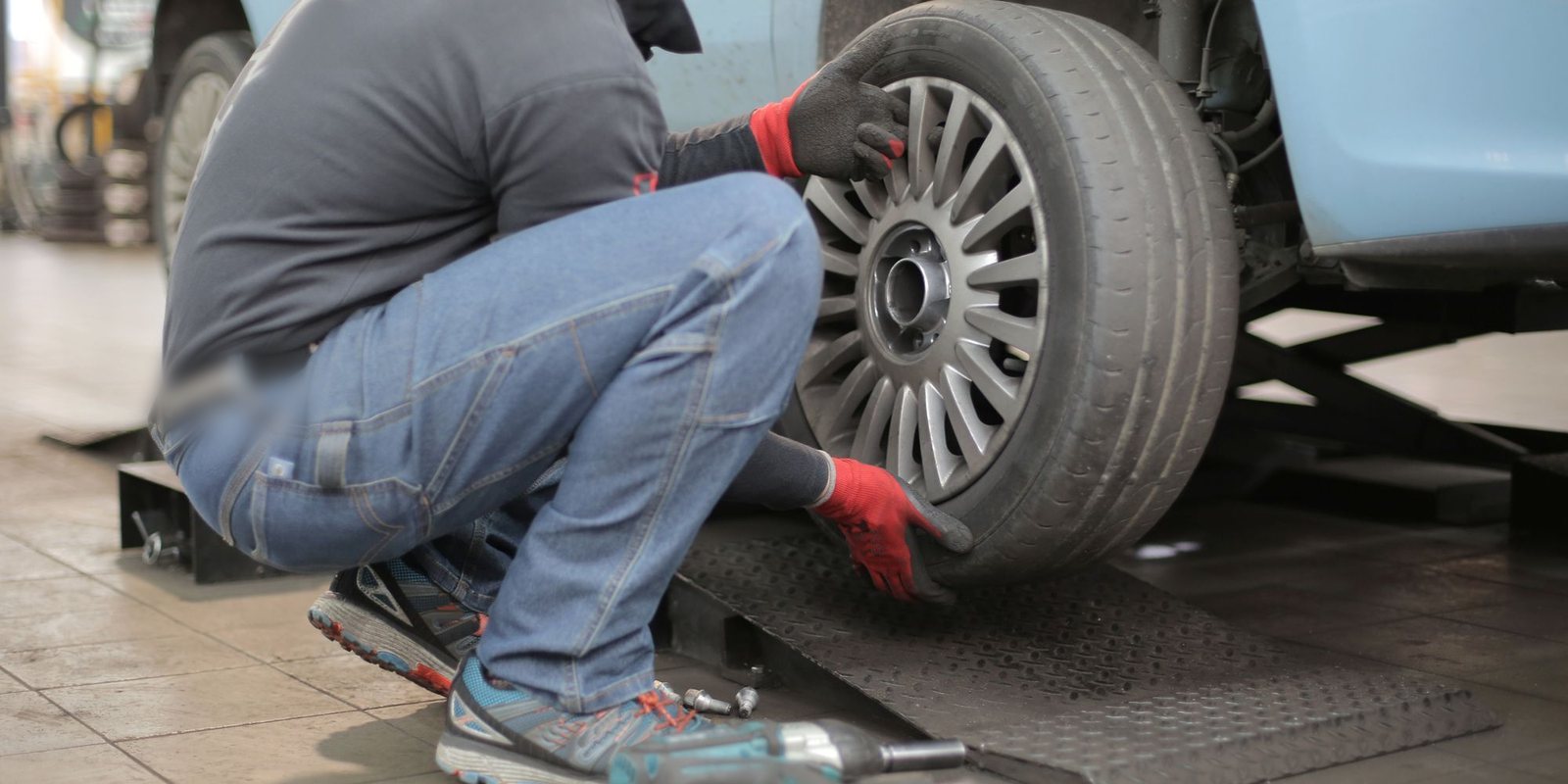 Técnico de luvas vermelhas agachado ao lado de um carro, com a mão apoiada no pneu e na roda, numa oficina de piso claro